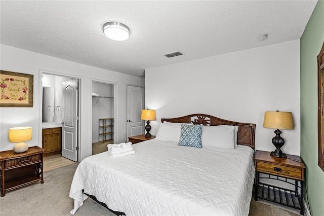 bedroom with visible vents, a walk in closet, light carpet, ensuite bathroom, and a textured ceiling