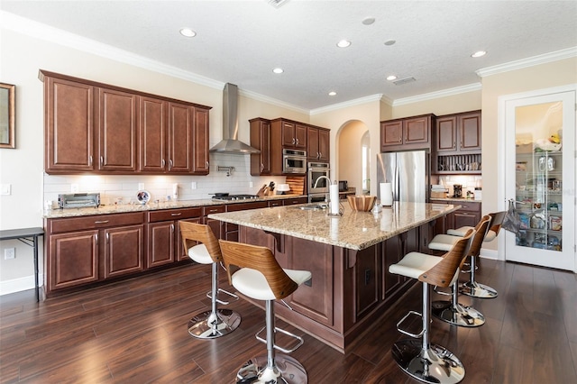 kitchen featuring dark wood-type flooring, arched walkways, stainless steel appliances, wall chimney exhaust hood, and a kitchen island with sink