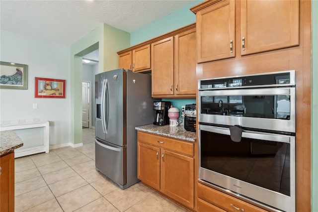 kitchen with light stone counters, a textured ceiling, appliances with stainless steel finishes, brown cabinetry, and light tile patterned floors