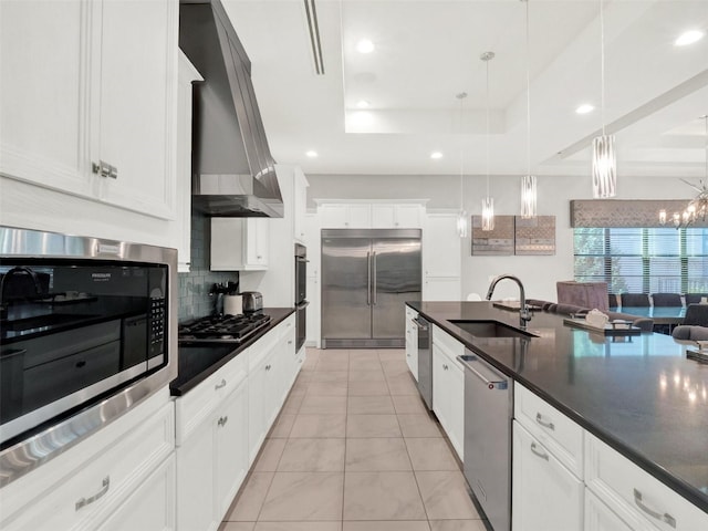 kitchen with dark countertops, wall chimney range hood, built in appliances, a raised ceiling, and a sink