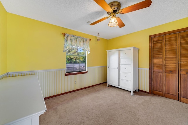 unfurnished bedroom with ceiling fan, light colored carpet, a wainscoted wall, and a textured ceiling