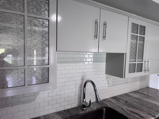 interior details featuring decorative backsplash, a sink, white cabinetry, dark stone counters, and glass insert cabinets