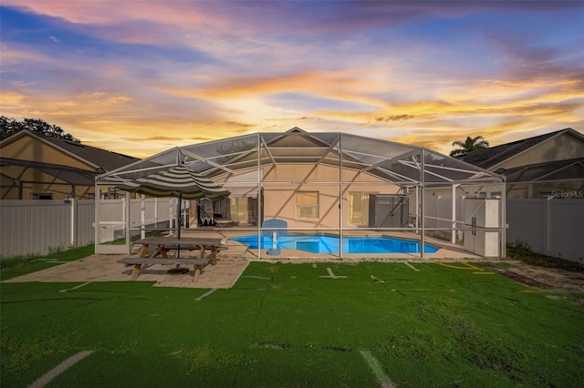 back of property at dusk featuring glass enclosure, a fenced in pool, a fenced backyard, a patio area, and a lawn