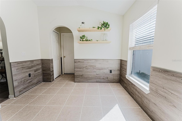 empty room featuring light tile patterned floors, lofted ceiling, arched walkways, wainscoting, and tile walls