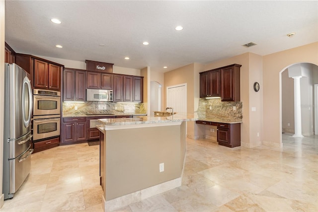 kitchen with a center island with sink, decorative columns, light stone counters, arched walkways, and stainless steel appliances