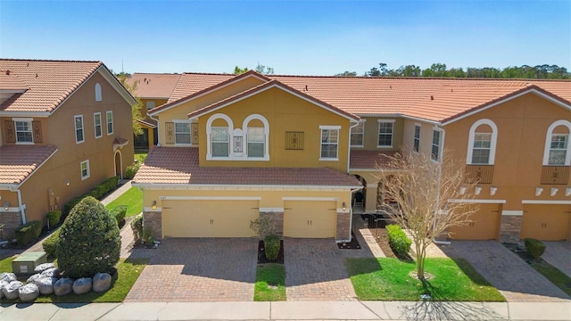 townhome / multi-family property with a tile roof, stucco siding, decorative driveway, stone siding, and an attached garage