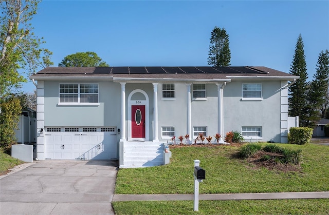 raised ranch with solar panels, a front yard, stucco siding, driveway, and an attached garage