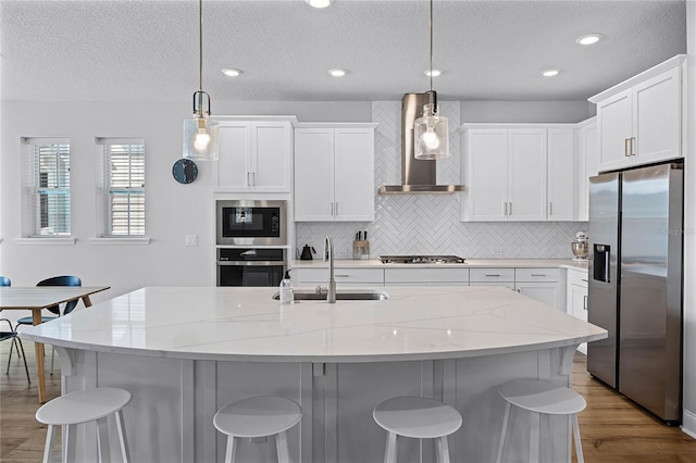 kitchen featuring wall chimney range hood, a sink, light wood-style floors, appliances with stainless steel finishes, and a kitchen breakfast bar