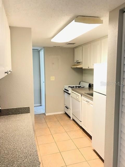 kitchen with white appliances, light tile patterned flooring, under cabinet range hood, a textured ceiling, and white cabinetry