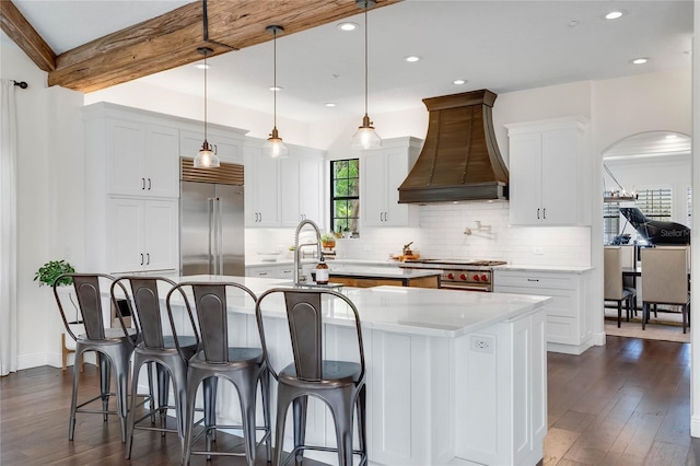 kitchen with high end appliances, a center island with sink, arched walkways, custom range hood, and beamed ceiling