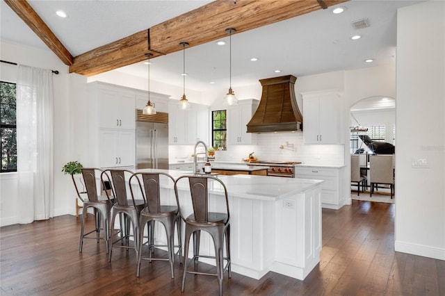 kitchen featuring beam ceiling, a center island with sink, custom range hood, premium appliances, and tasteful backsplash