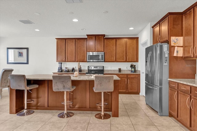 kitchen featuring a kitchen island, light stone countertops, light tile patterned floors, brown cabinetry, and stainless steel appliances