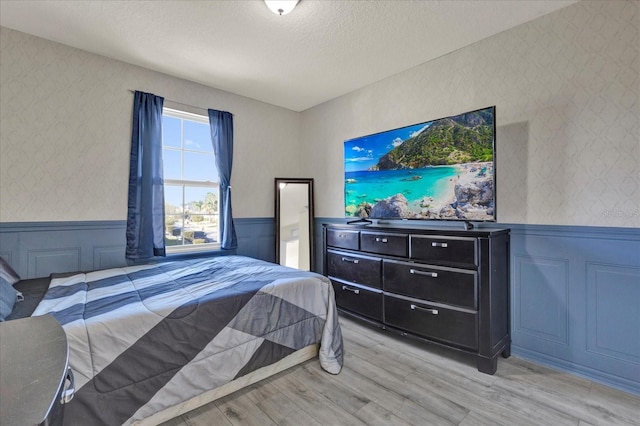 bedroom featuring wainscoting, a textured ceiling, wallpapered walls, and light wood-style floors