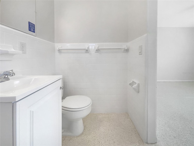 half bath featuring tile walls, a wainscoted wall, toilet, vanity, and speckled floor