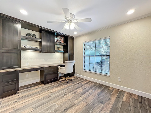 unfurnished office featuring ceiling fan, baseboards, light wood-type flooring, built in desk, and a textured wall