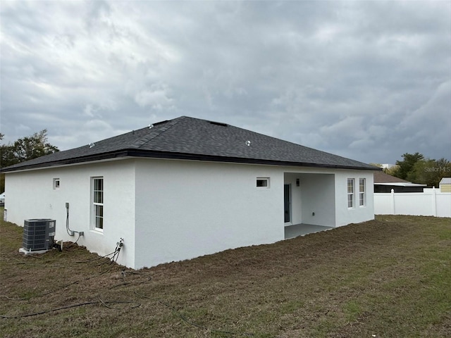 back of house featuring stucco siding, a lawn, central AC, fence, and roof with shingles