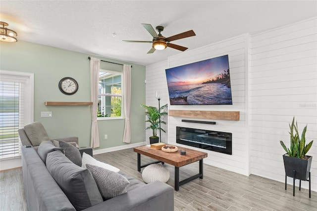 living area featuring a textured ceiling, wood finished floors, a glass covered fireplace, baseboards, and ceiling fan