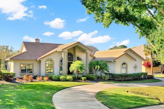 mediterranean / spanish-style home featuring curved driveway, a front lawn, a tiled roof, stucco siding, and a chimney