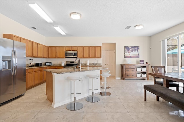 kitchen with stone counters, brown cabinets, appliances with stainless steel finishes, a kitchen breakfast bar, and a kitchen island with sink