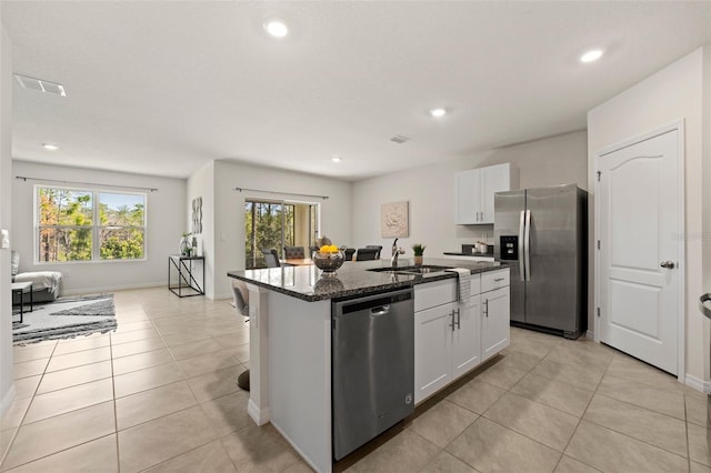 kitchen with light tile patterned floors, a center island with sink, dark stone counters, a sink, and stainless steel appliances