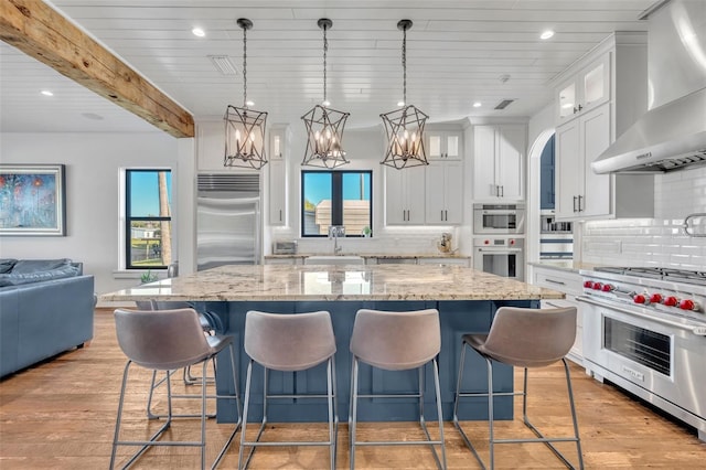 kitchen with high end appliances, wall chimney exhaust hood, glass insert cabinets, and plenty of natural light