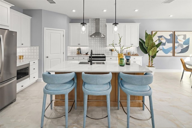 kitchen with an island with sink, a sink, appliances with stainless steel finishes, a kitchen bar, and wall chimney range hood