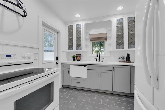 kitchen featuring glass insert cabinets, light countertops, gray cabinets, white appliances, and a sink