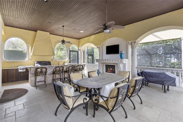 view of patio with grilling area, a lit fireplace, outdoor dining area, an outdoor kitchen, and a ceiling fan