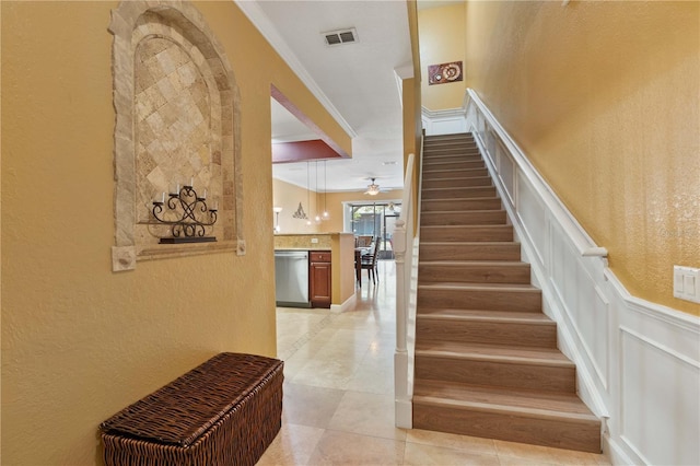 stairway featuring a decorative wall, ceiling fan, visible vents, and ornamental molding