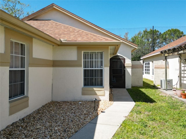 doorway to property featuring stucco siding, central AC unit, a yard, and roof with shingles