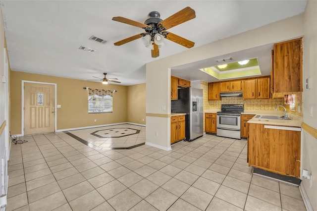 kitchen featuring a tray ceiling, a sink, stainless steel appliances, under cabinet range hood, and brown cabinets