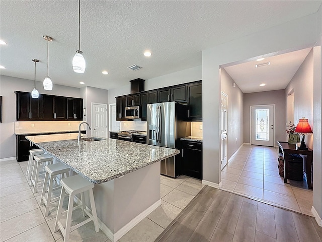 kitchen featuring a sink, dark cabinetry, appliances with stainless steel finishes, decorative backsplash, and a large island with sink