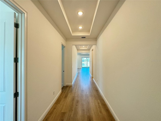 hallway with a tray ceiling, visible vents, baseboards, and light wood finished floors