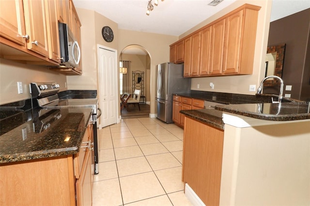 kitchen with dark stone countertops, stainless steel appliances, arched walkways, a peninsula, and light tile patterned floors