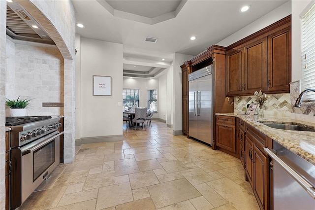 kitchen with a sink, premium appliances, stone tile flooring, wall chimney exhaust hood, and a raised ceiling