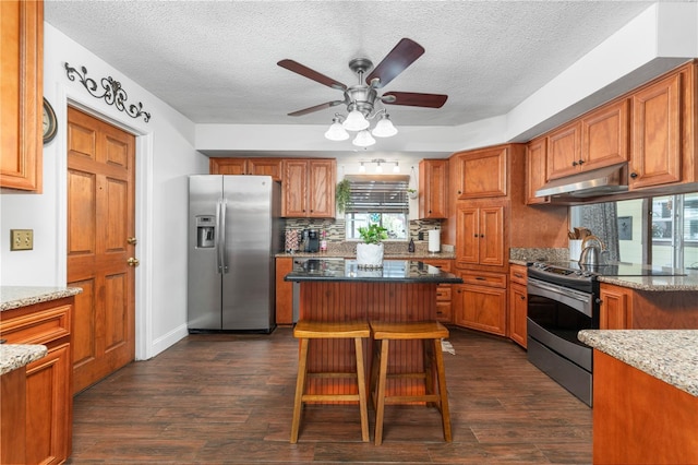 kitchen featuring light stone counters, a ceiling fan, stainless steel appliances, under cabinet range hood, and brown cabinets