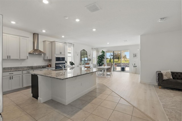 kitchen featuring light stone countertops, visible vents, a kitchen island with sink, built in microwave, and wall chimney range hood