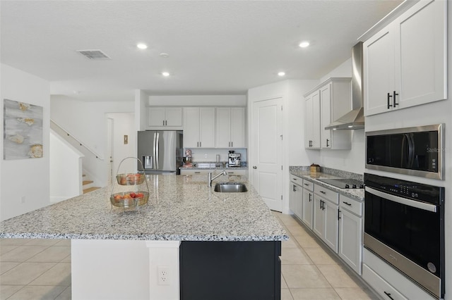 kitchen featuring an island with sink, appliances with stainless steel finishes, light tile patterned flooring, wall chimney exhaust hood, and a sink