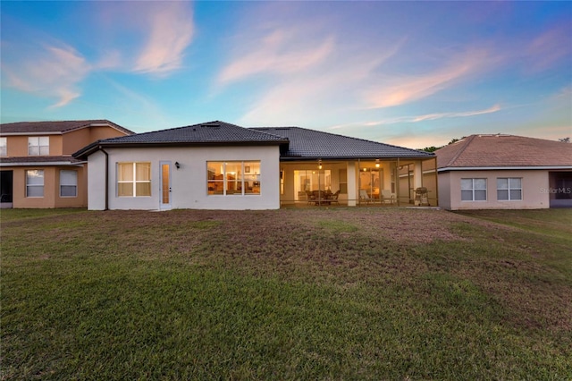 back of house at dusk featuring a tile roof, a lawn, and stucco siding