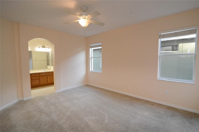 unfurnished bedroom featuring ensuite bathroom, a ceiling fan, arched walkways, baseboards, and light colored carpet