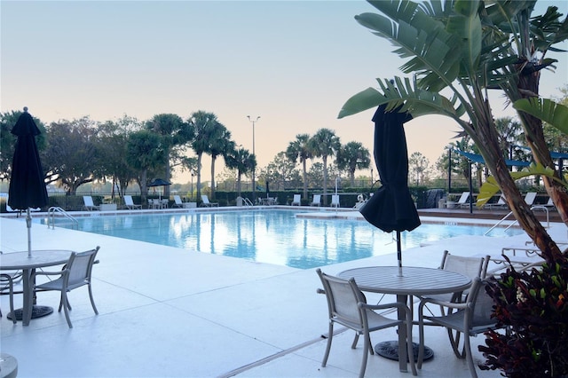 pool at dusk with a patio area, outdoor dining area, fence, and a community pool