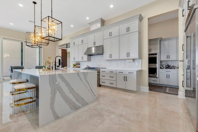 kitchen with light stone countertops, appliances with stainless steel finishes, under cabinet range hood, marble finish floor, and a chandelier