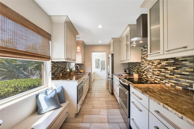 kitchen with a sink, dark stone countertops, stainless steel appliances, wall chimney range hood, and glass insert cabinets