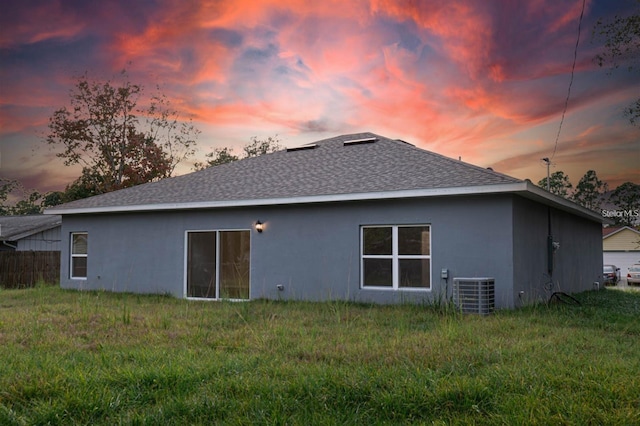 back of property at dusk featuring a shingled roof, cooling unit, a yard, and stucco siding