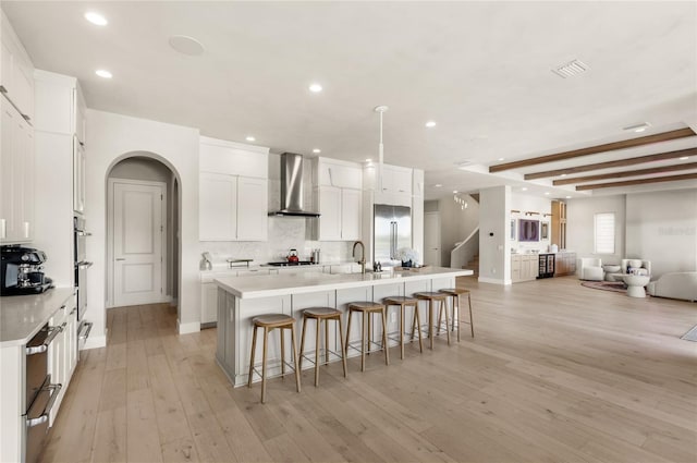 kitchen with stainless steel appliances, open floor plan, light wood-style floors, wall chimney exhaust hood, and tasteful backsplash