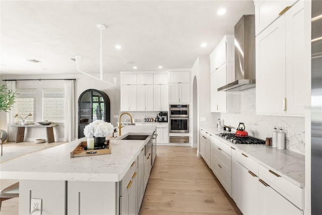 kitchen featuring a sink, backsplash, light wood-style floors, appliances with stainless steel finishes, and wall chimney exhaust hood