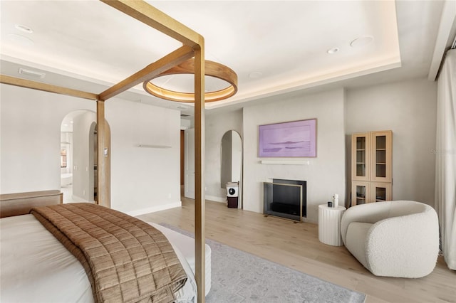 bedroom with light wood-style floors, a raised ceiling, a fireplace, and arched walkways