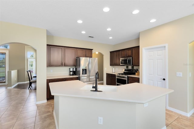 kitchen featuring dark brown cabinetry, an island with sink, light countertops, arched walkways, and stainless steel appliances