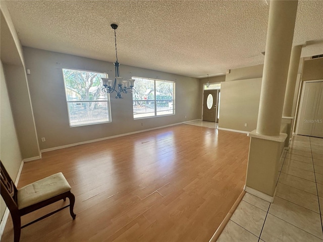 unfurnished dining area with light wood-style flooring, a textured ceiling, baseboards, a chandelier, and ornate columns