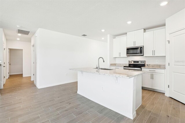 kitchen featuring a sink, a kitchen island with sink, light wood finished floors, appliances with stainless steel finishes, and light stone countertops
